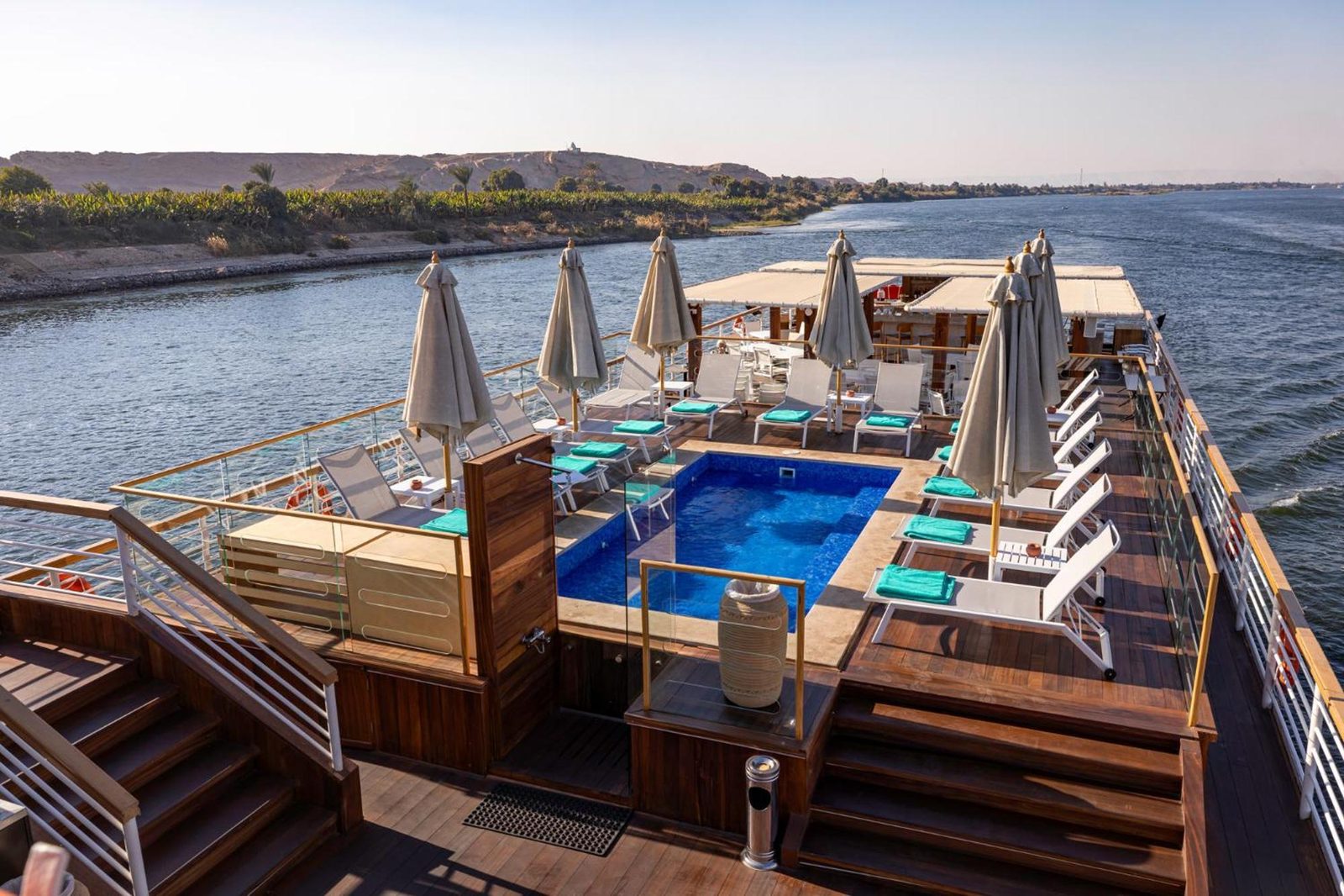 The teak sundeck of the MS TIA with a plunge pool, loungers and shade umbrellas, the banks of the Nile sliding past